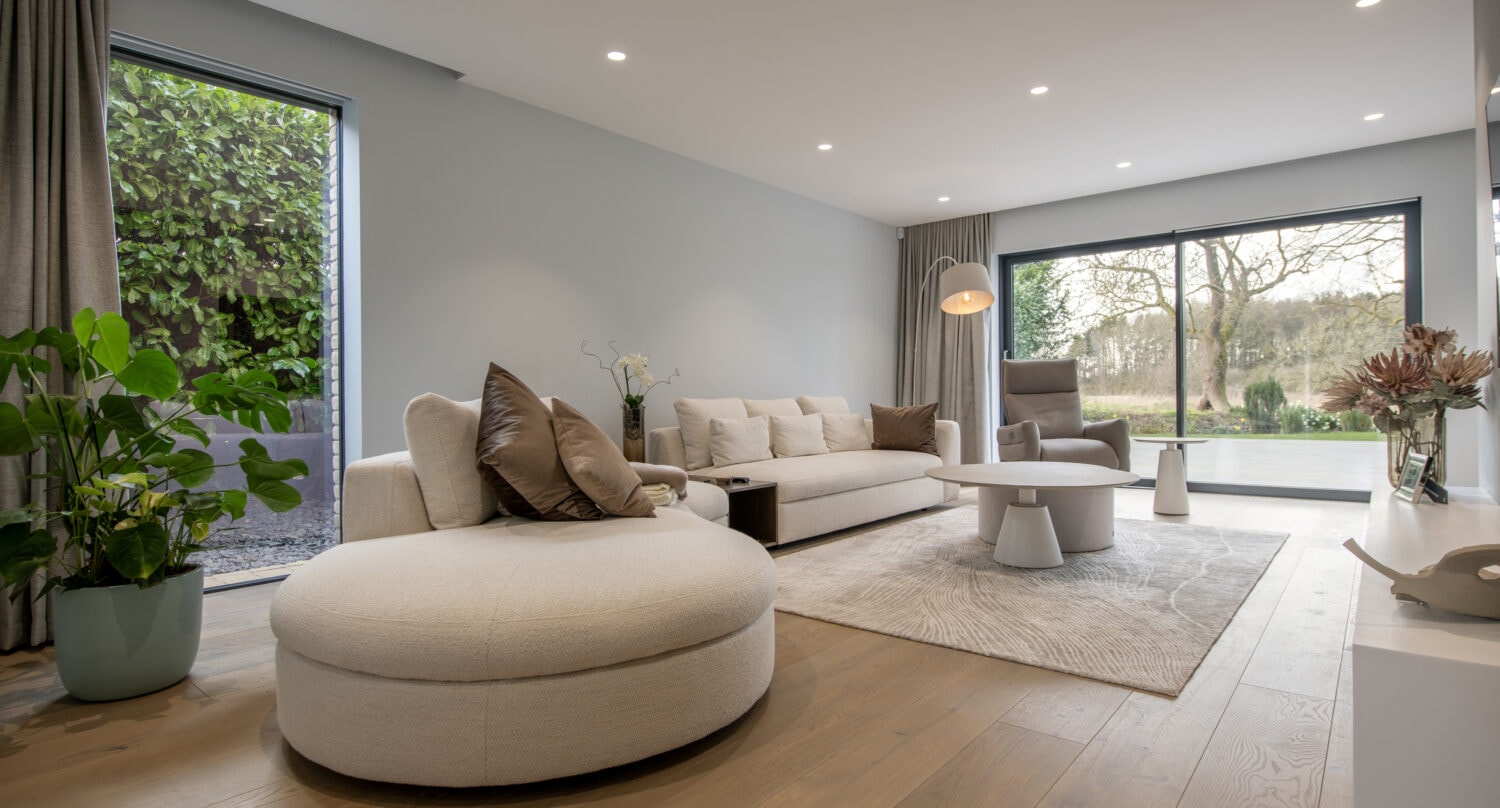 Modern living room with large windows, neutral-toned furniture, a round chair, sofa with cushions, coffee table, light wood flooring, indoor plants, and bifold doors offering a stunning view of the green garden outside.