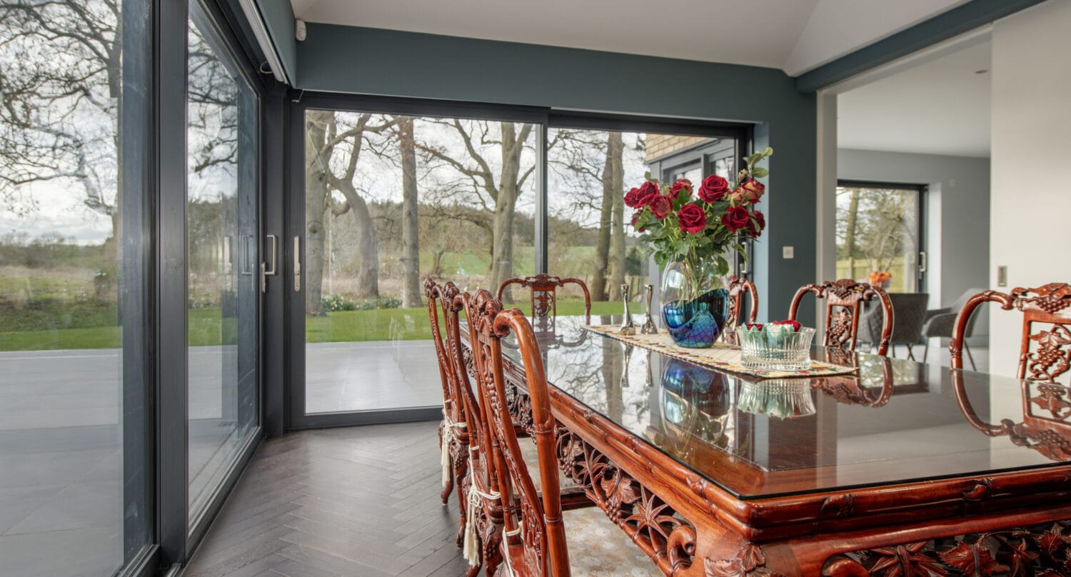 A dining room with carved wooden chairs around a polished table, a vase of red roses as centerpiece, and large bifold doors offering a view of trees and greenery outside.