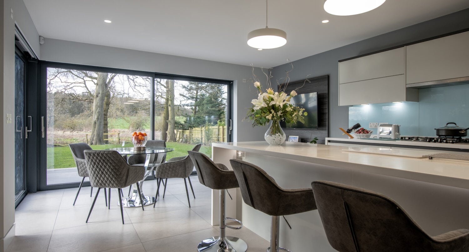Modern kitchen with a breakfast bar and three gray stools, a round dining table with four chairs by bespoke glass sliding doors, and a view of a green garden outside; contemporary lighting and fresh flower arrangements are present.