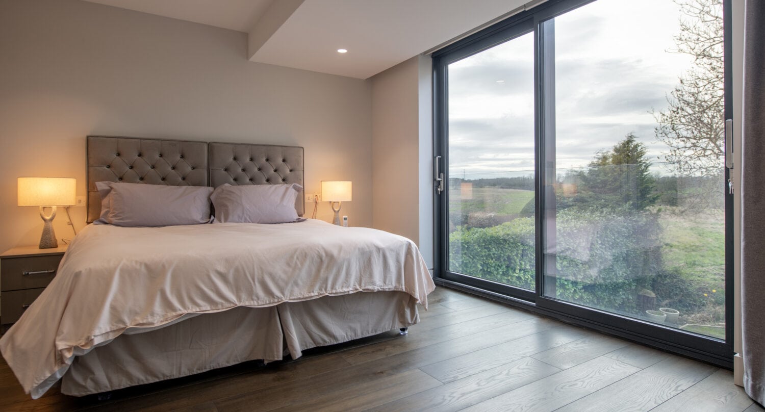 A modern bedroom with a large bed, two bedside tables with lamps, wooden floor, and floor-to-ceiling sliding doors offering a view of trees and open landscape outside.