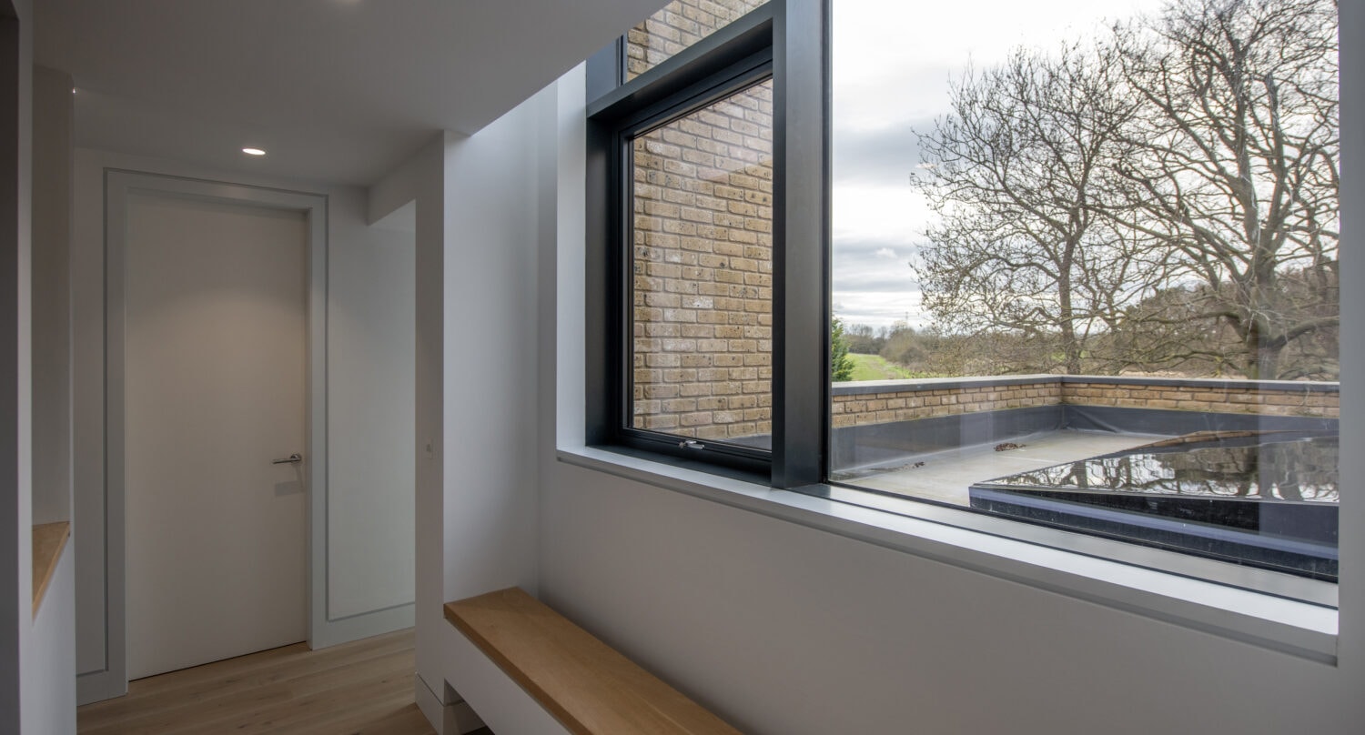 A modern hallway with light wooden floors, a wooden bench along the window, and a door at the end. Large windows and bespoke glass solutions reveal a view of a brick wall, leafless tree, and cloudy sky outside.