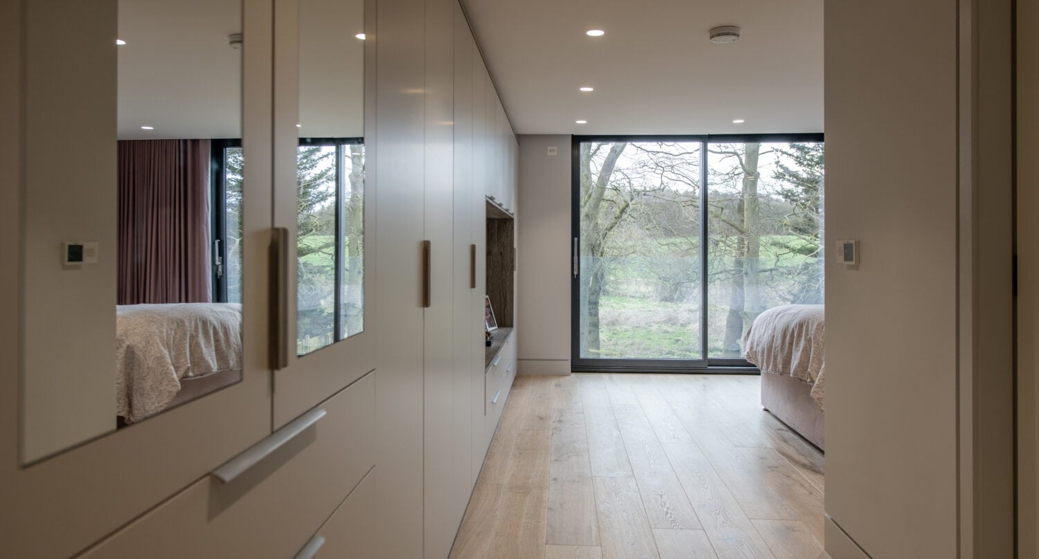 A modern bedroom with light wood floors, built-in wardrobes with bifold doors, and large floor-to-ceiling windows overlooking a green landscape. A bed is partially visible on the right side of the room.