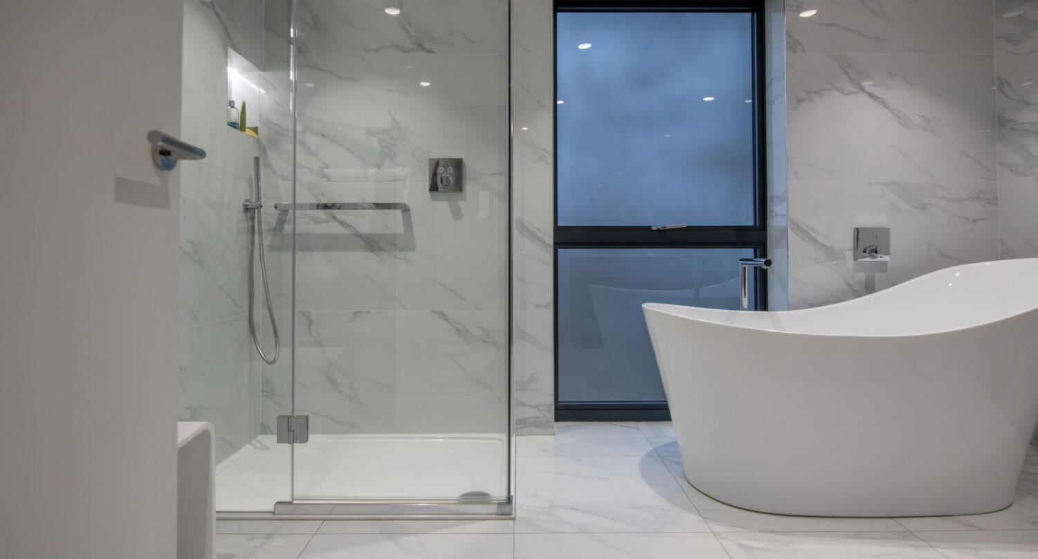 Modern bathroom with white marble walls and floor, a glass-enclosed shower on the left, bespoke glass solutions in design details, a freestanding white bathtub on the right, and a large window in the center background.