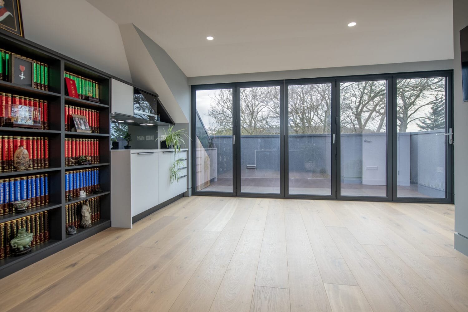 Modern room with light wood flooring, bespoke glass solutions featuring a wall of glass doors opening to a balcony, built-in kitchen area, and bookshelf filled with colorful encyclopedias and décor. Trees are visible outside through the glass.