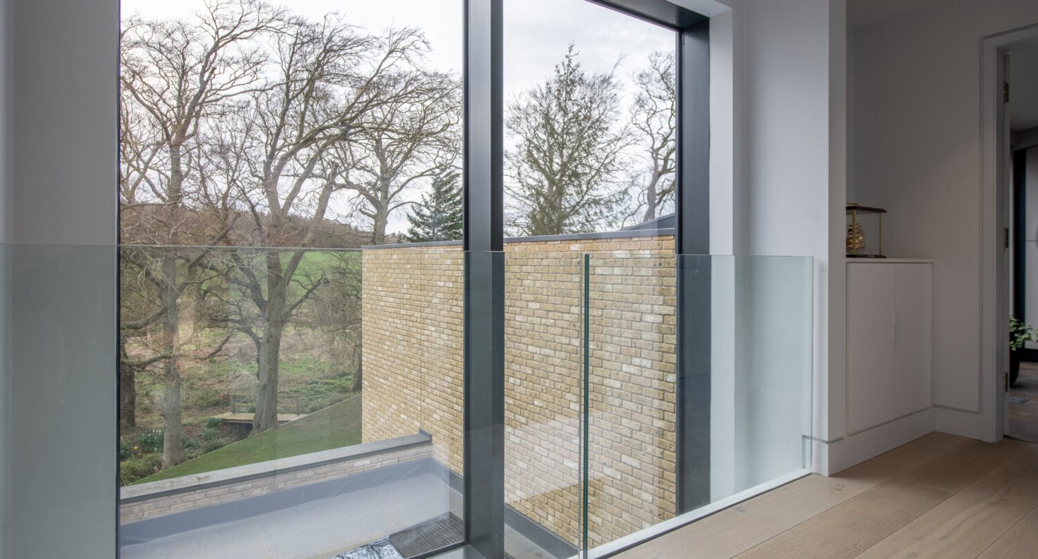 Floor-to-ceiling windows and bespoke glass solutions overlook trees and a brick wall in this modern home. Light wood flooring, white walls, and a glass railing line the edge near the window for an airy, contemporary feel.