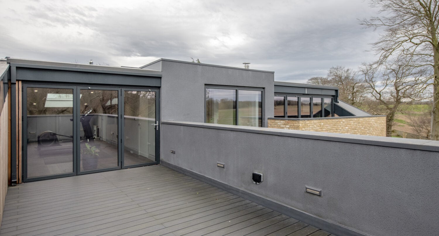 A modern rooftop terrace with gray decking and walls, large bifold doors, and windows. The terrace overlooks trees and a partly cloudy sky.