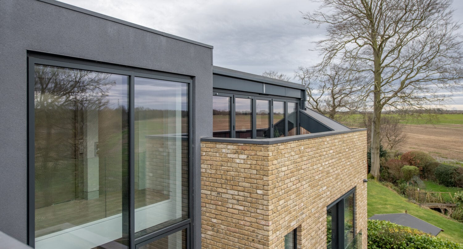 Modern house with large glass windows and yellow brick exterior, featuring bespoke glass solutions like bifold doors and a balcony, surrounded by trees and open fields under a cloudy sky.