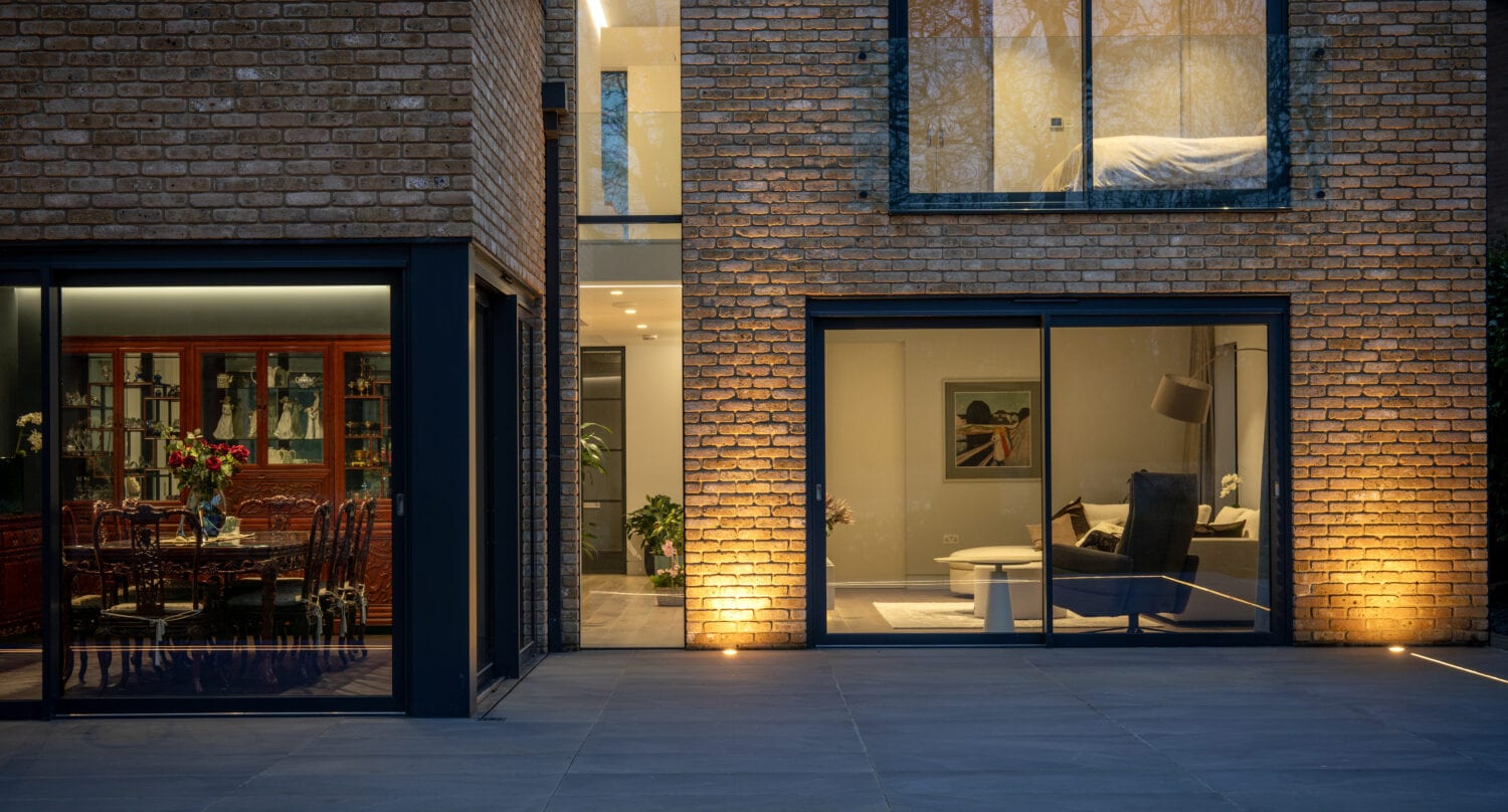 Modern brick house exterior at dusk with large bifold doors revealing a dining room with a table and chairs on the left and a cozy living room with an armchair and coffee table on the right, illuminated by warm lighting.