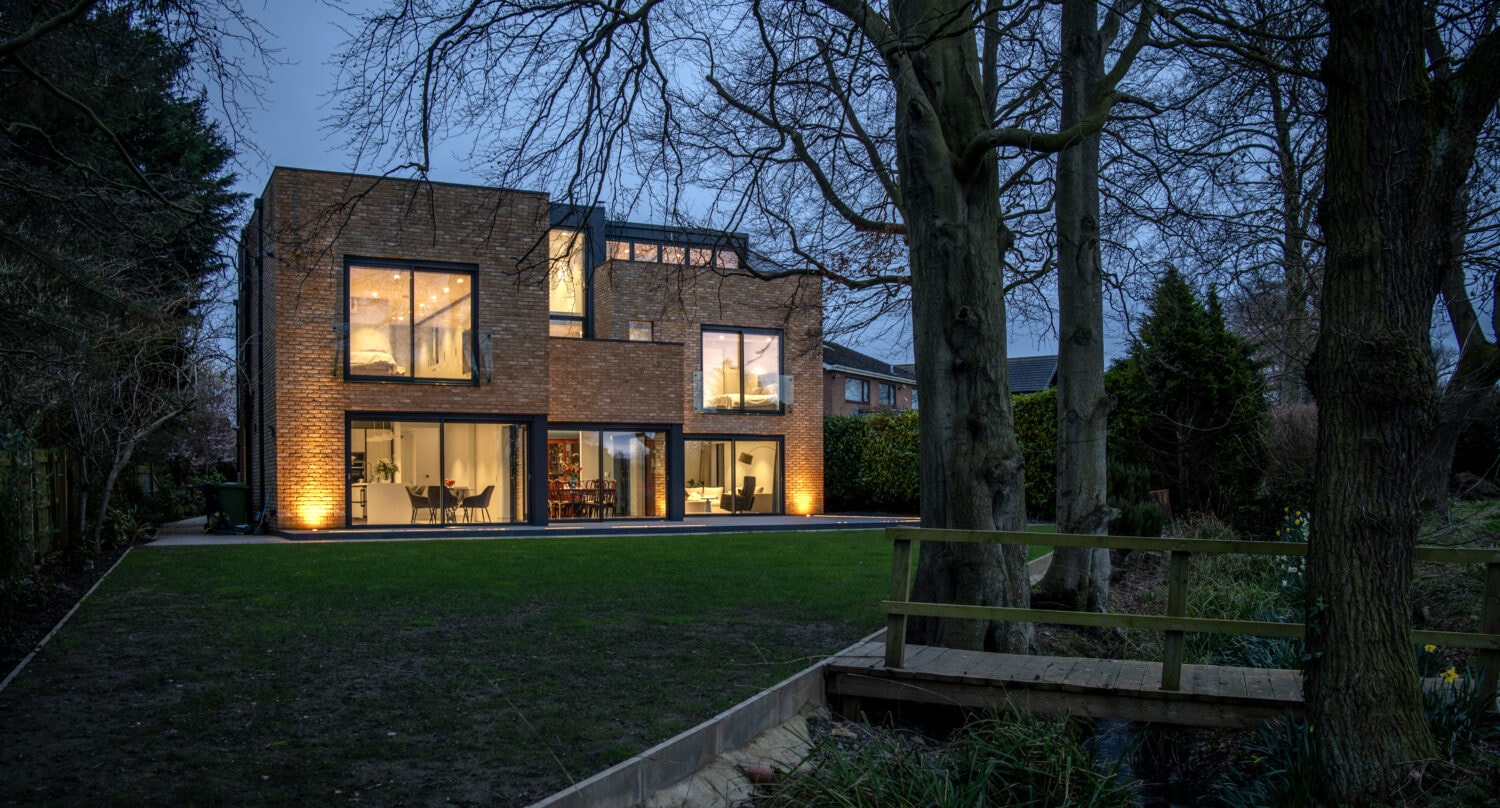 A modern two-story house with large, illuminated windows features bespoke glass solutions and sliding doors, sitting in a yard with a lawn, trees, and a small wooden bridge over a stream at dusk.