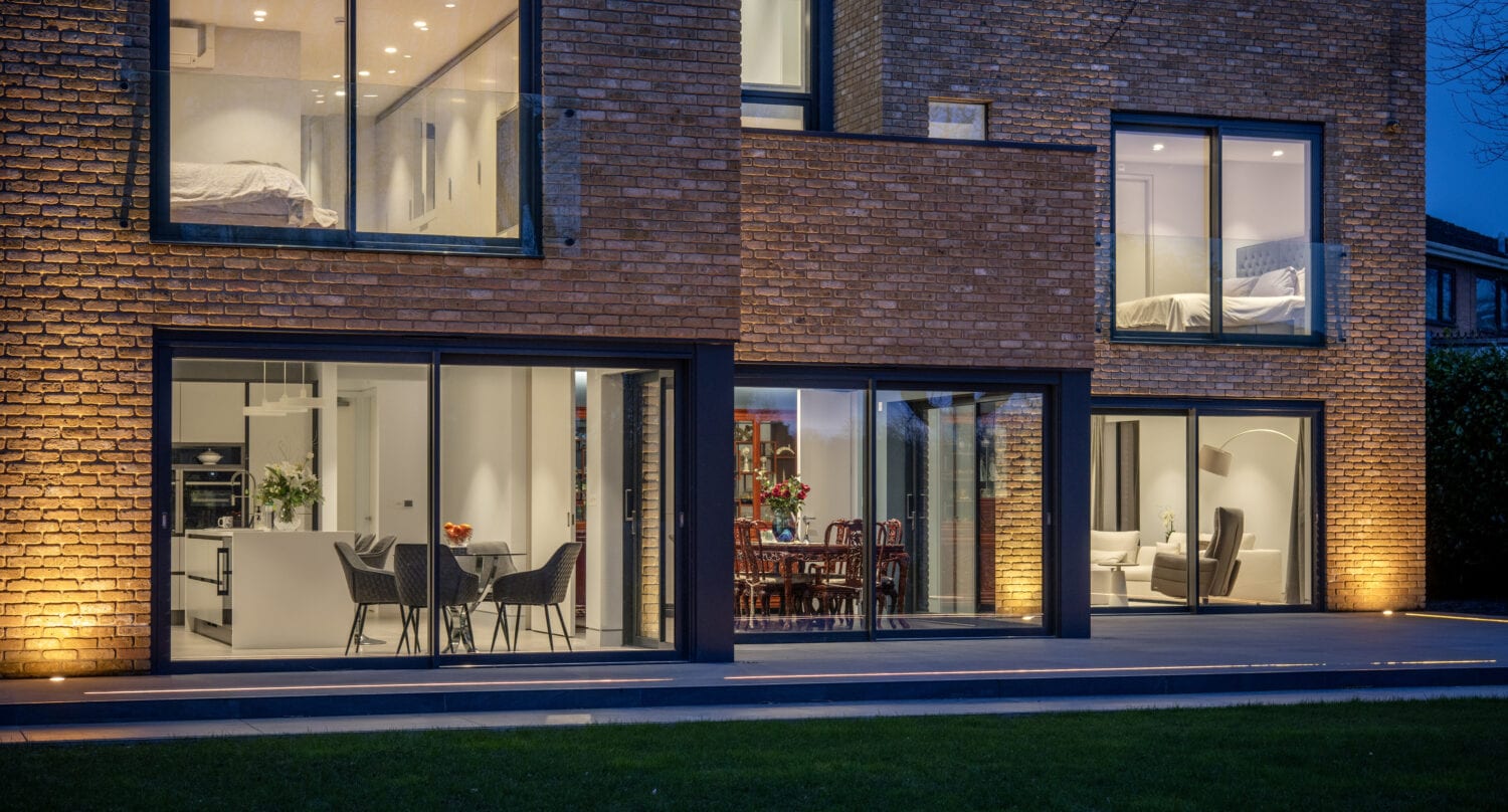 Modern brick house at dusk, with large glass windows and stylish bifold doors revealing well-lit interior spaces including dining and living areas. Bedrooms on the upper floor and exterior lights illuminate the facade.