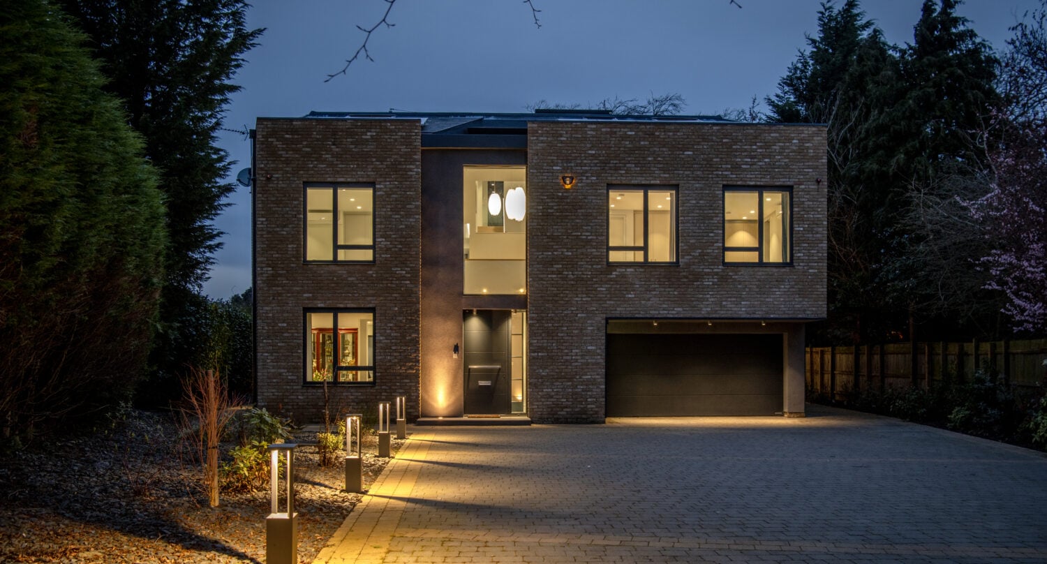 Modern two-story house with large windows, brick exterior, and a double garage, featuring bespoke glass solutions like sliding doors. Warm outdoor lights illuminate a paved driveway at dusk, surrounded by trees and landscaped garden beds.