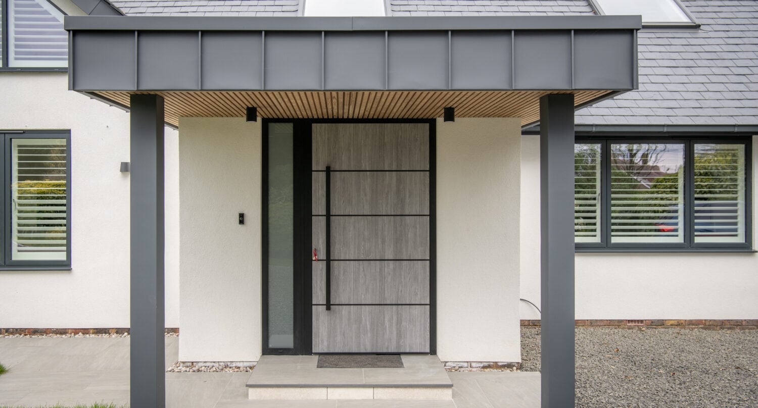 Modern house entrance with a wide, light wood front door, large sidelights, and a gray overhang supported by two pillars. White walls and horizontal windows with shutters frame the front doors, while gravel and grass complete the inviting look.