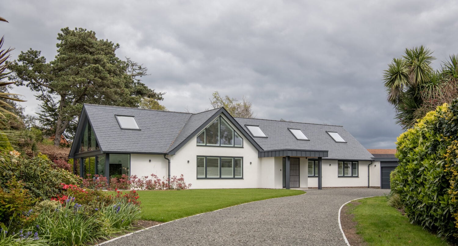 Modern single-story house with large windows, sleek glass roofing, and a dark gray sloped roof, surrounded by a well-kept lawn, colorful flowers, and trees under a cloudy sky. A gravel driveway leads up to the entrance.