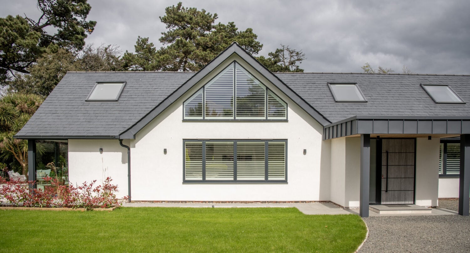 Modern white house with dark gray roof, skylights, large triangular window above main window, and a covered entrance. Bespoke glass solutions like bifold doors open to a well-maintained green lawn and landscaped garden. Trees and cloudy sky in the background.