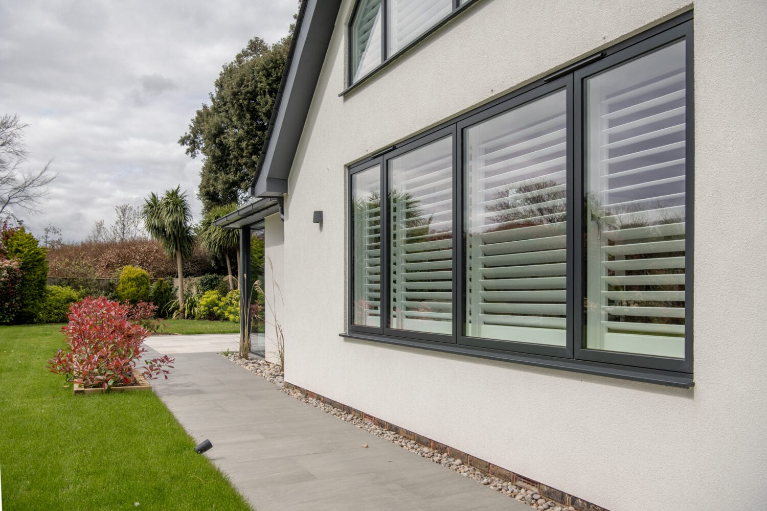 Modern house exterior with large windows featuring white plantation shutters, light-colored walls, a paved walkway, green lawn, landscaping with shrubs and trees, and stylish bifold doors under a cloudy sky.