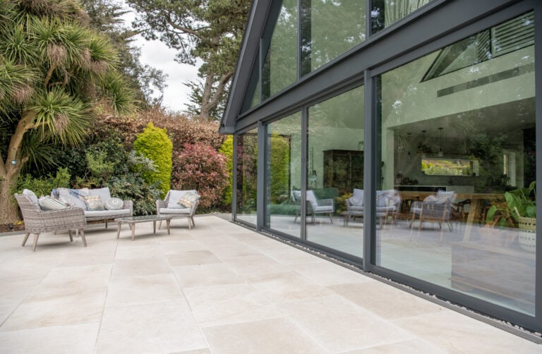 A modern patio with light stone tiles features outdoor seating and is bordered by lush greenery. Bespoke glass solutions, including large sliding doors, seamlessly connect the patio to a stylish, open-plan indoor living space.