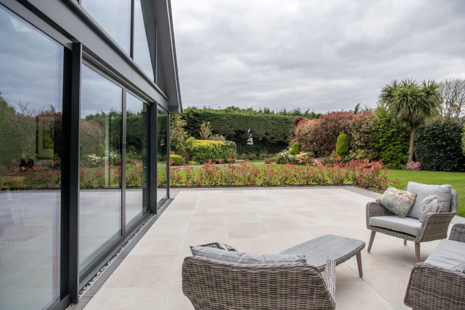 A modern patio with wicker chairs and a small table overlooks a landscaped garden. Large sliding doors reflect the vibrant flowers, green shrubs, and trees under a cloudy sky, seamlessly connecting indoor comfort with outdoor beauty.