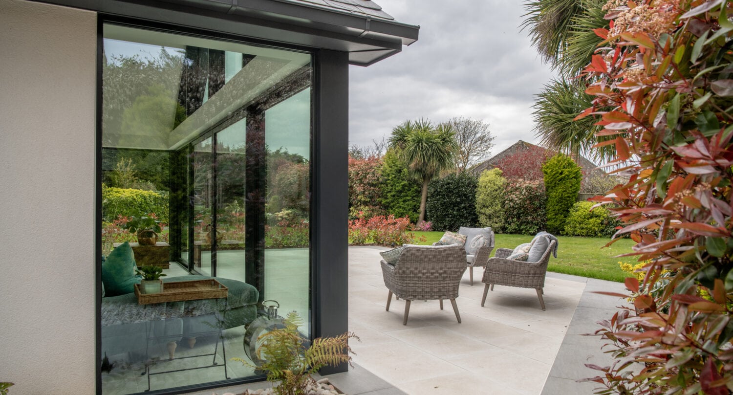 Modern patio with wicker chairs and a table on a tiled terrace, next to large bespoke glass solutions windows. The area is surrounded by lush green garden, palm trees, and colorful shrubs under a cloudy sky.