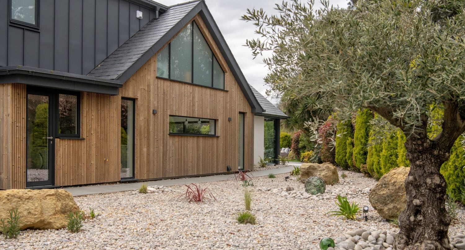 Modern house with wooden and black panel exterior, large windows, bespoke glass solutions, and a minimalist front yard landscaped with rocks, small plants, and a few trees under a cloudy sky.