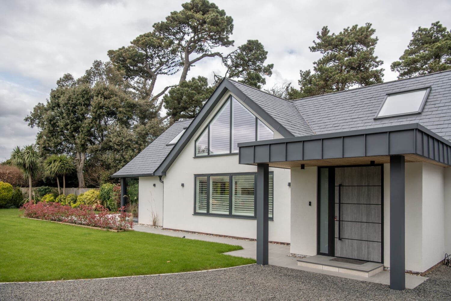 Modern single-story house with white walls, large windows, and a gray sloped roof. Featuring bifold doors that open to a manicured lawn, with shrubs and tall trees in the background under a cloudy sky.