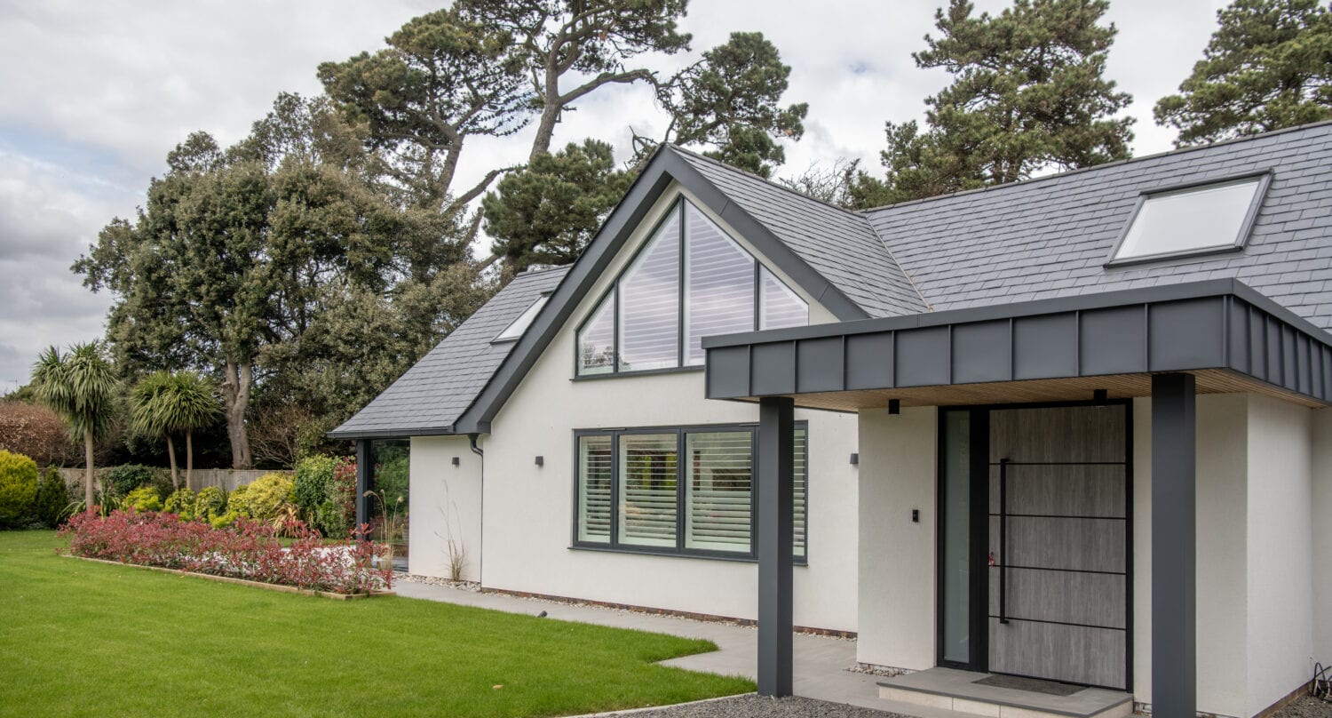Modern single-story house with white walls, large windows, and a gray sloped roof. Featuring bifold doors that open to a manicured lawn, with shrubs and tall trees in the background under a cloudy sky.