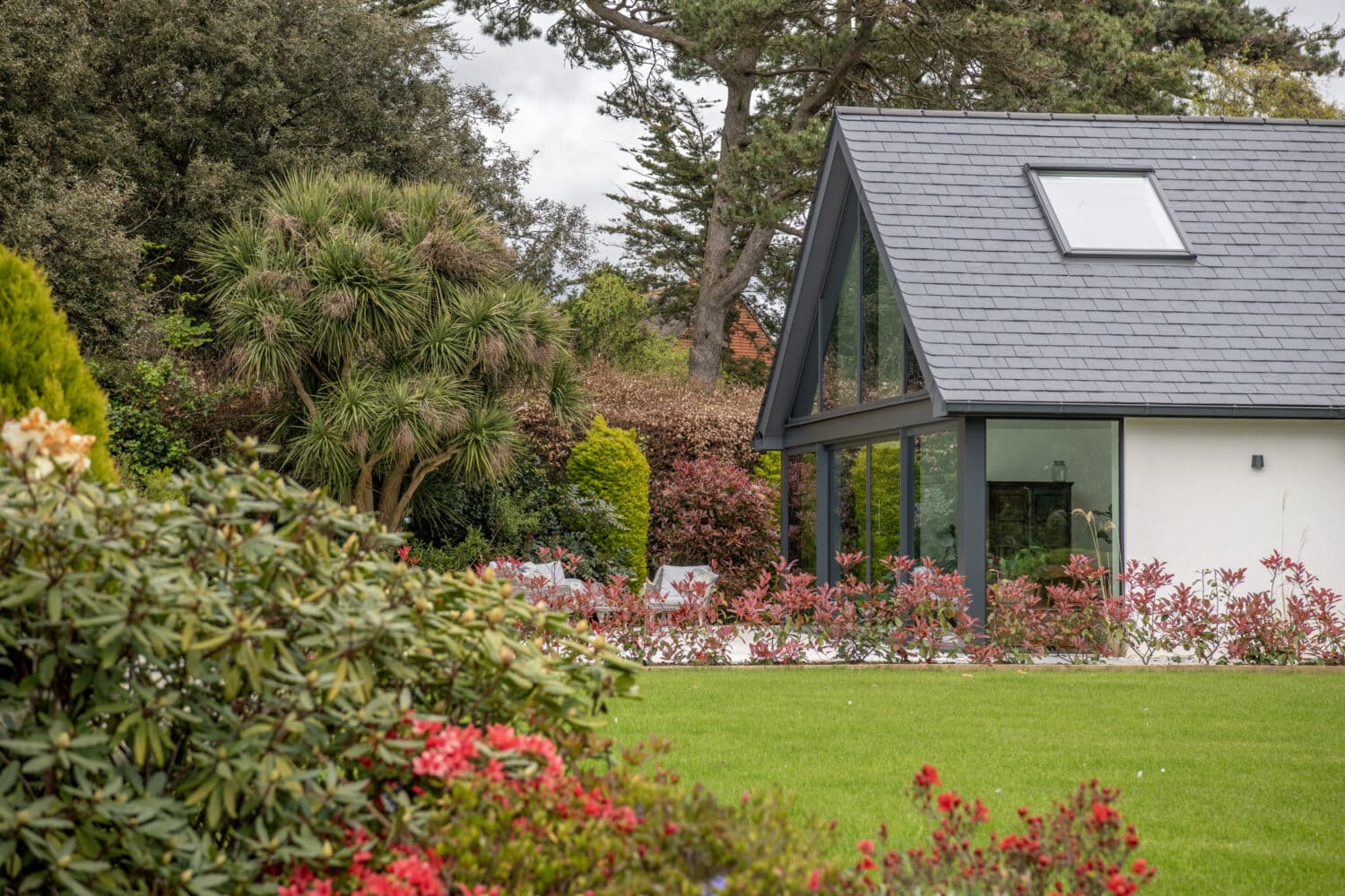 A modern house with large glass windows and bespoke glass solutions sits beside a lush, green garden filled with vibrant red and pink flowers and various shrubs, surrounded by tall trees under a cloudy sky.