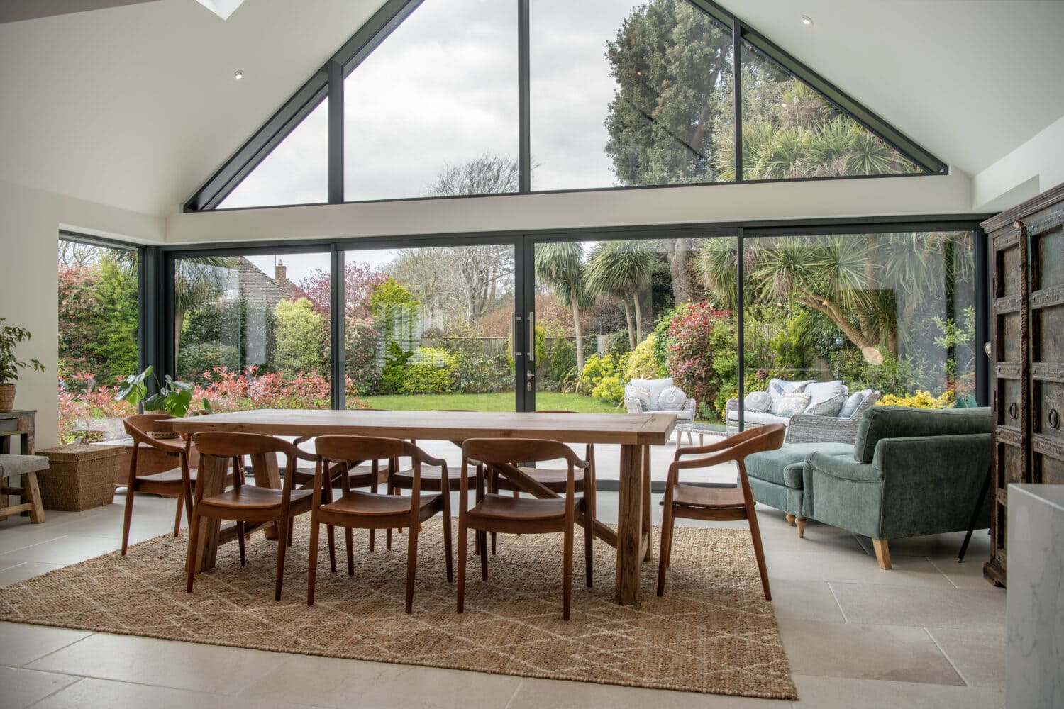 A modern dining area with a wooden table and chairs sits on a woven rug. Large bifold doors open to a lush garden, letting in natural light. A green sofa and outdoor seating are visible through the windows.