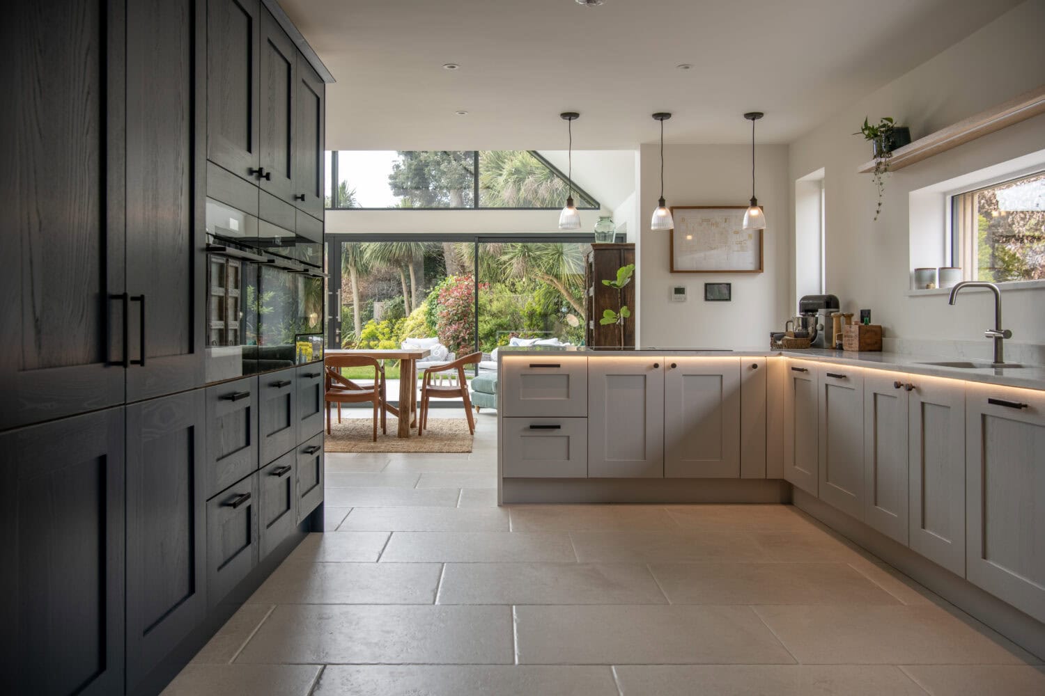 Modern kitchen with grey cabinets, large tile flooring, pendant lights, and a spacious dining area featuring bifold doors opening to a lush, green garden. Natural light fills the room through bespoke glass solutions and large windows.