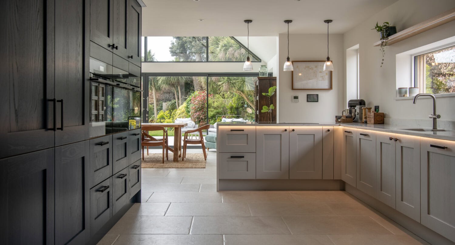 Modern kitchen with grey cabinets, large tile flooring, pendant lights, and a spacious dining area featuring bifold doors opening to a lush, green garden. Natural light fills the room through bespoke glass solutions and large windows.