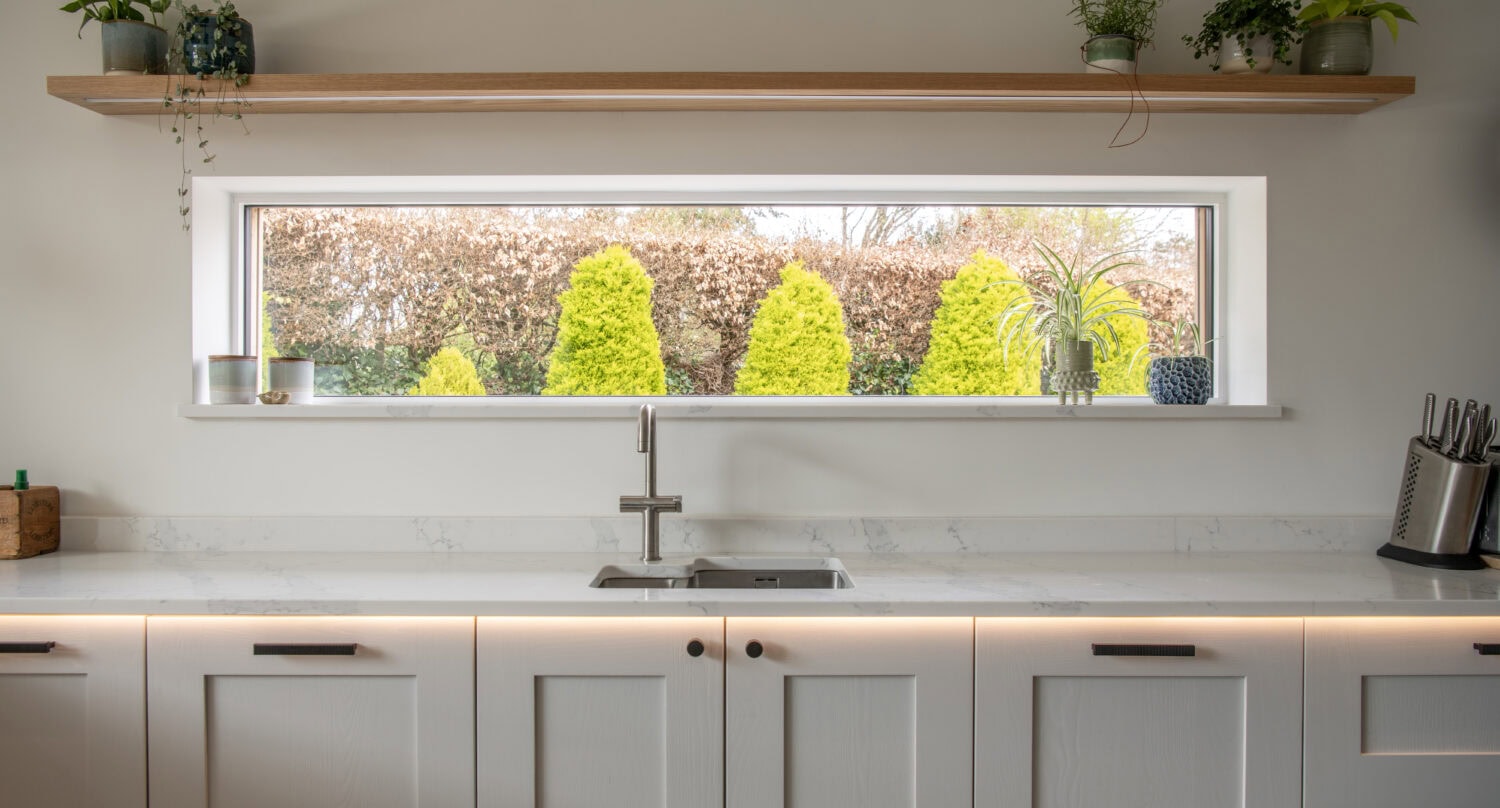 Modern kitchen with white cabinets, a sleek sink, and a window overlooking a garden with bright green shrubs. Bespoke glass solutions above the window enhance natural light, while potted plants add vibrant greenery inside.