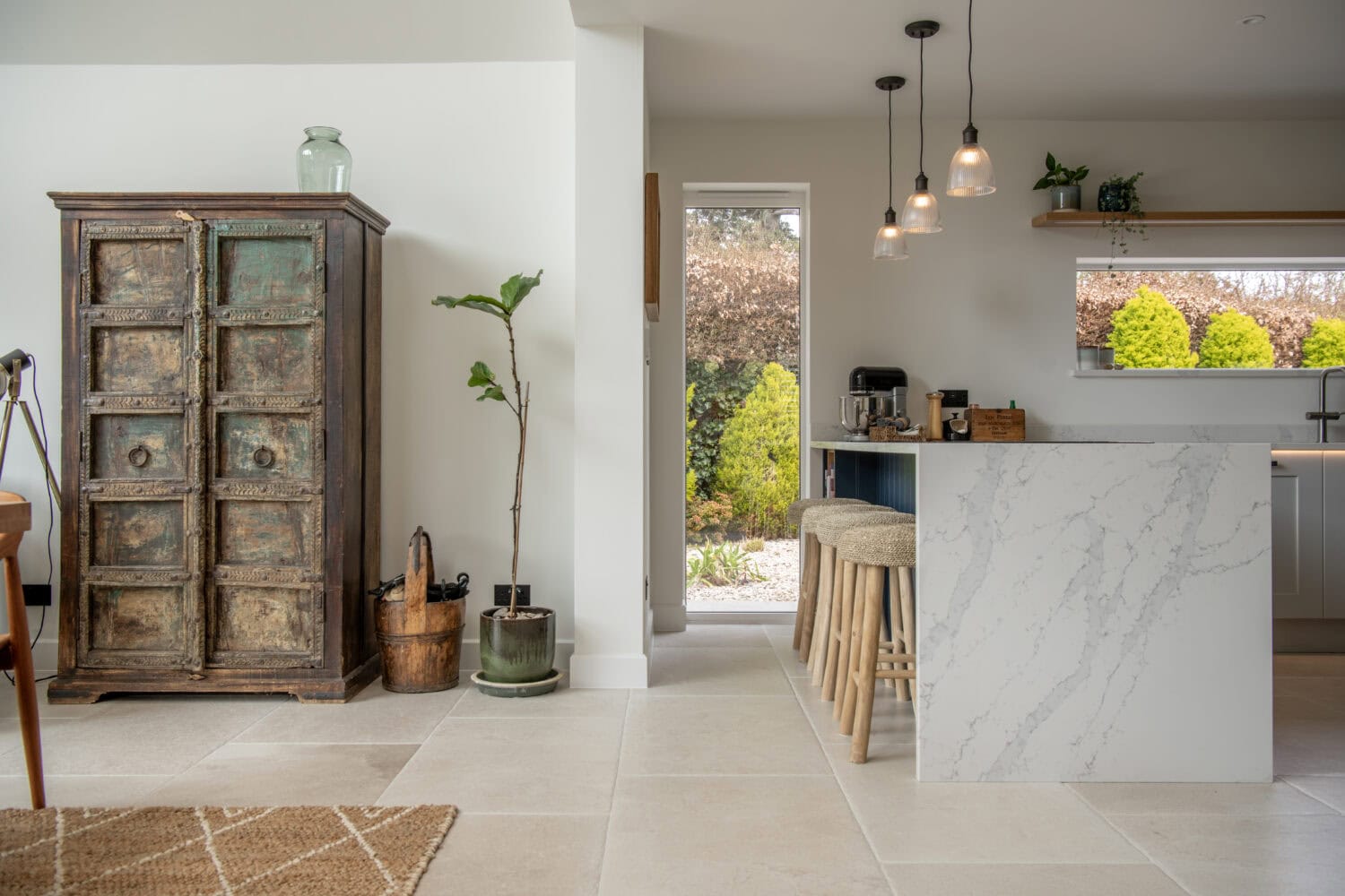 A bright, modern kitchen with a marble island, wooden stools, hanging pendant lights, a rustic cabinet, potted plants, and bespoke glass solutions like sliding doors leading to a sunny garden with green hedges.