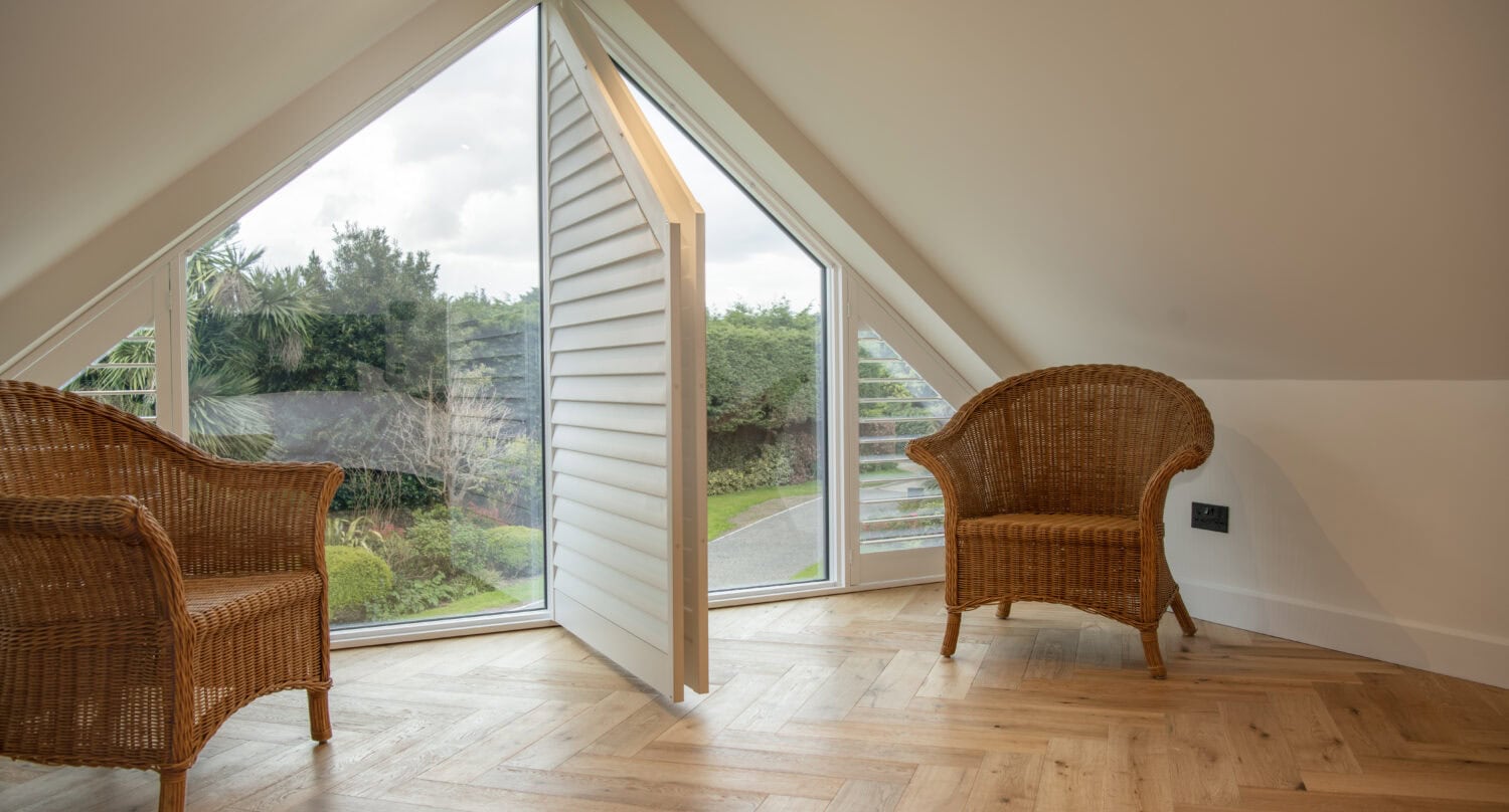 A bright attic room with wooden herringbone flooring, two wicker chairs, and a triangular window with white plantation shutters, complemented by bespoke glass solutions overlooking a green garden.