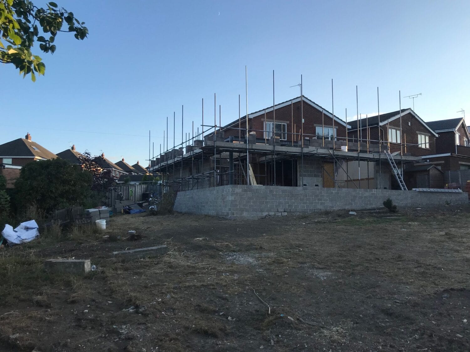A house under construction with scaffolding around the upper level, unfinished brickwork, and building materials scattered on the ground in front, featuring space for bifold doors, with neighboring homes visible in the background.