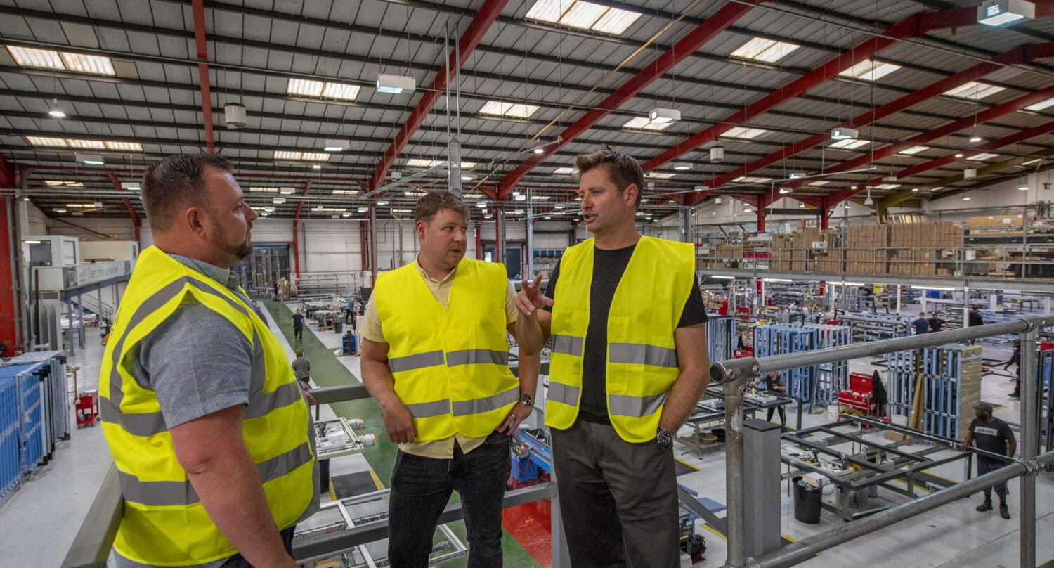 Three men wearing yellow safety vests stand and talk on a platform overlooking a large, busy industrial factory with metal structures, bright lights, and glass roofing.