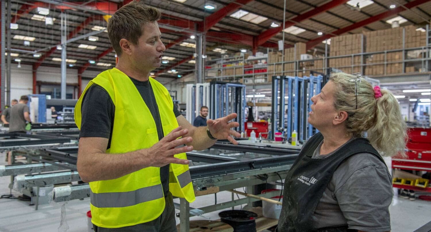 A man in a yellow safety vest talks to a woman in work overalls inside a large factory specializing in bespoke glass solutions, with machinery and other workers visible in the background.