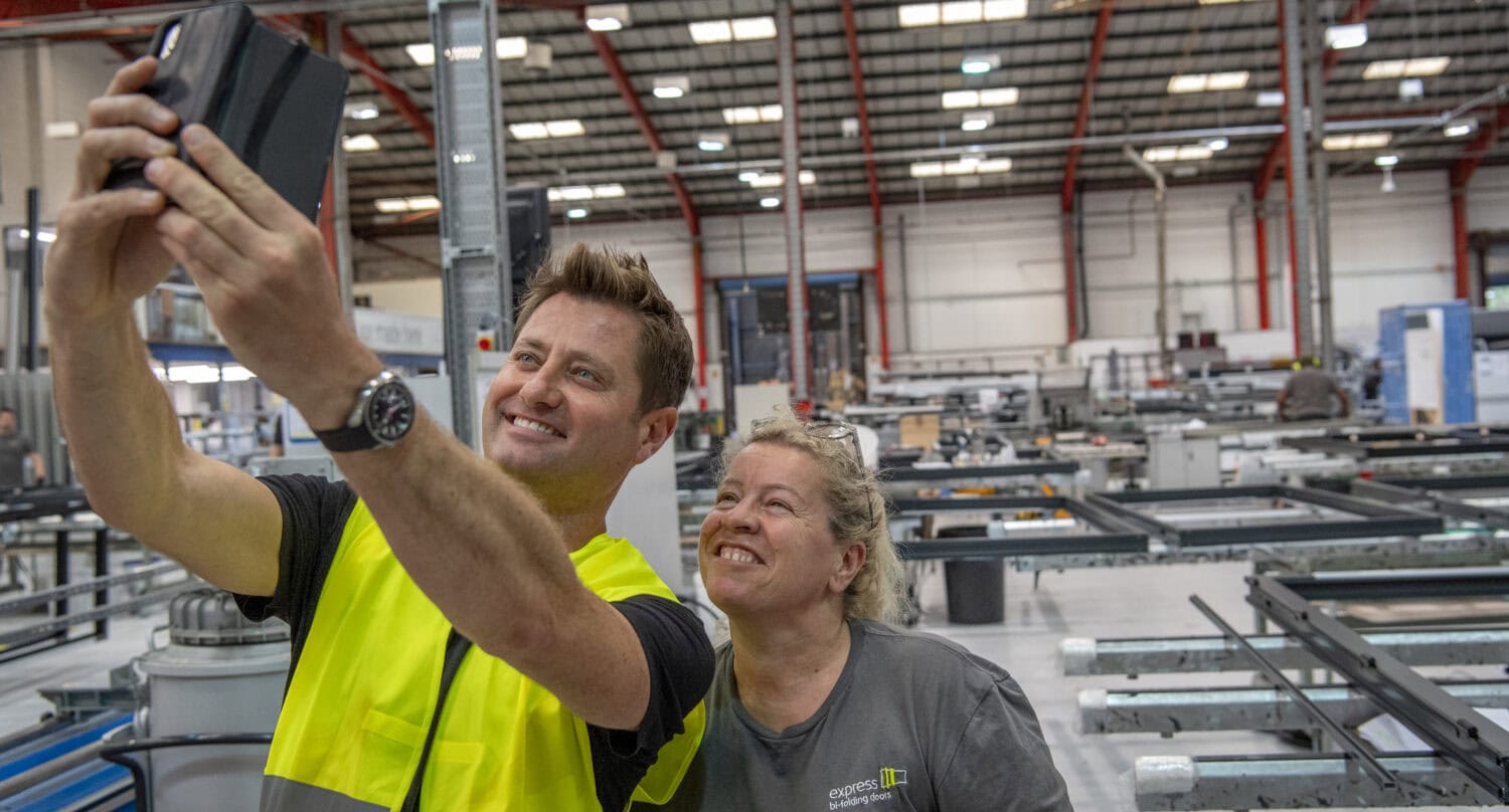 Two people in work attire smile and take a selfie together inside a large industrial factory, with machinery, equipment, and sleek bifold doors visible in the background.