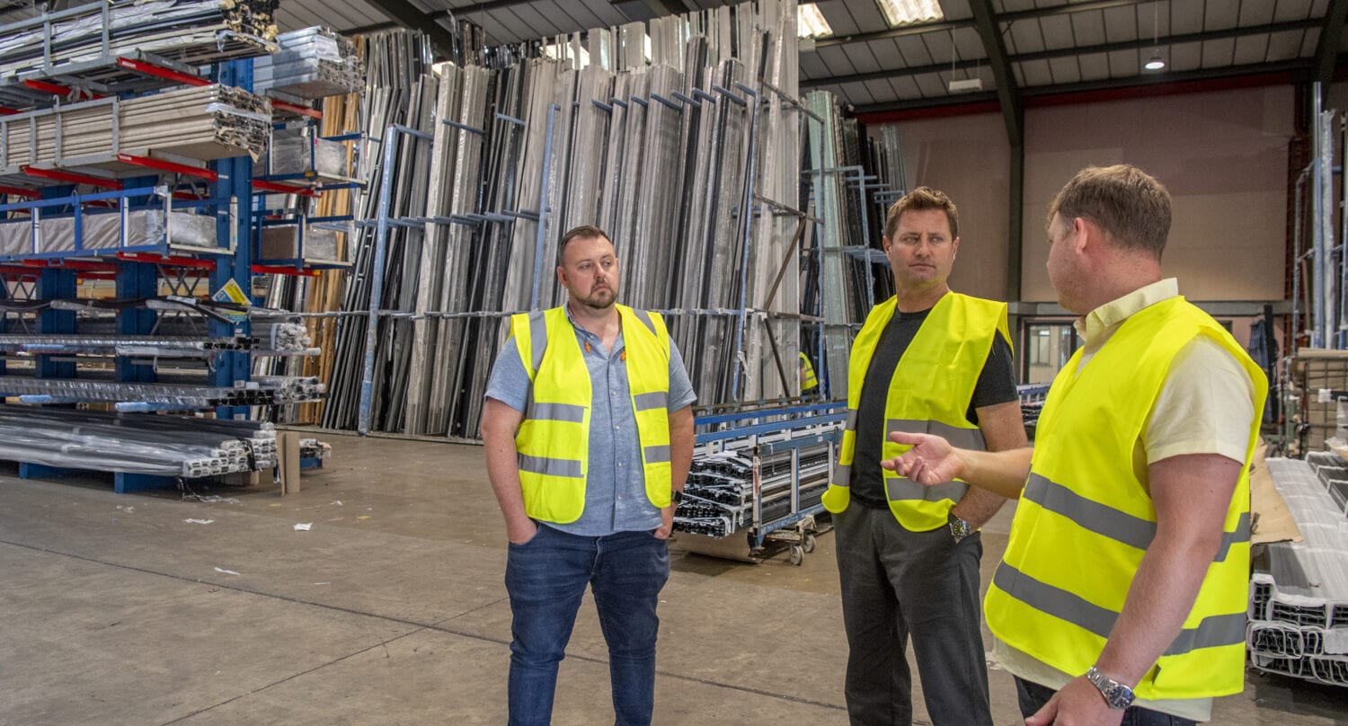 Three men wearing yellow safety vests stand and talk inside a large warehouse filled with stacked metal pipes and beams. The spacious area features high ceilings, industrial shelving, and sliding doors for easy access.