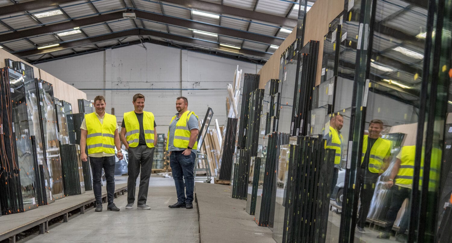 Three men in yellow safety vests stand in a warehouse aisle lined with large glass panes and materials for bespoke glass solutions, under a high ceiling with glass roofing and skylights.