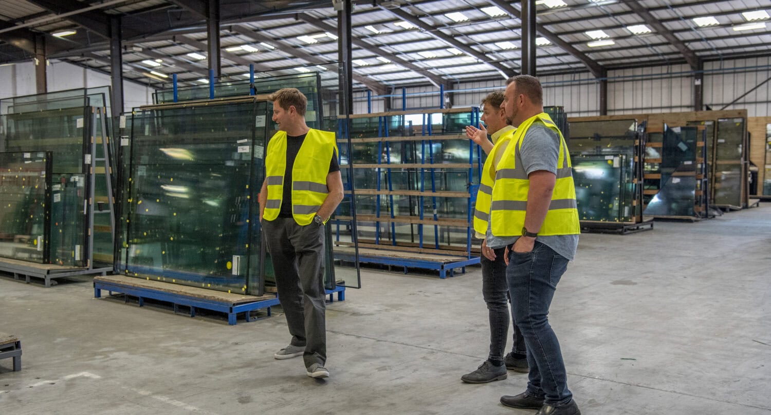 Three people wearing yellow safety vests walk through a spacious, well-lit warehouse filled with large glass panels for sliding doors on racks, with one person gesturing as they talk.
