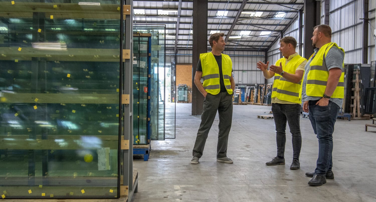 Three men wearing yellow safety vests stand and talk in a spacious warehouse, surrounded by large glass panels, industrial equipment, and bespoke glass solutions for sliding doors.