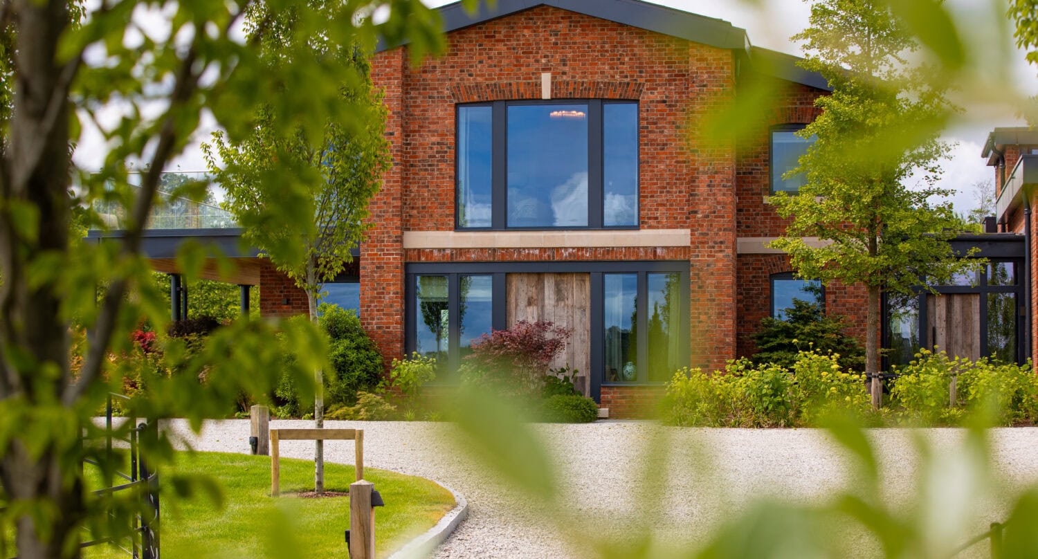 A modern brick house with large glass windows and bespoke glass solutions, surrounded by green trees and shrubs, is viewed through leafy branches and a curved gravel driveway in the foreground.