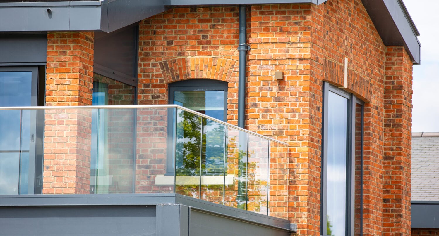 Modern brick house exterior with large glass windows, glass roofing, and a glass balcony railing, featuring a mix of traditional red brickwork and contemporary architectural elements.