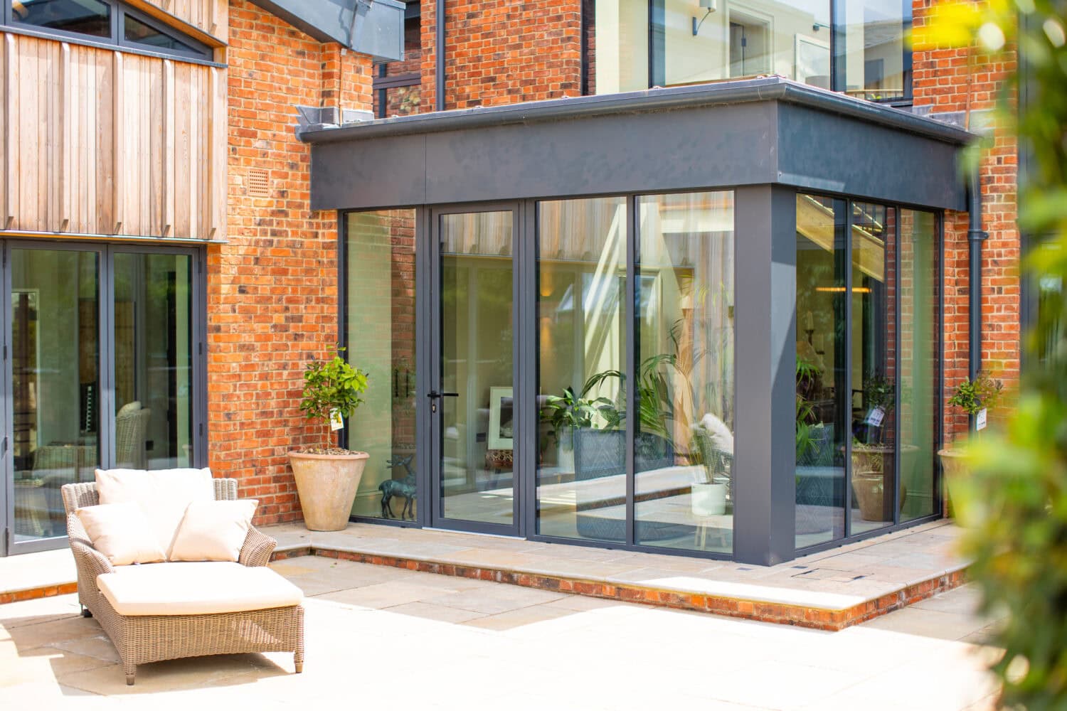 Modern patio area with a cushioned lounge chair in front of a house featuring large glass windows, bifold doors, red brick walls, and potted plants near the entrance.