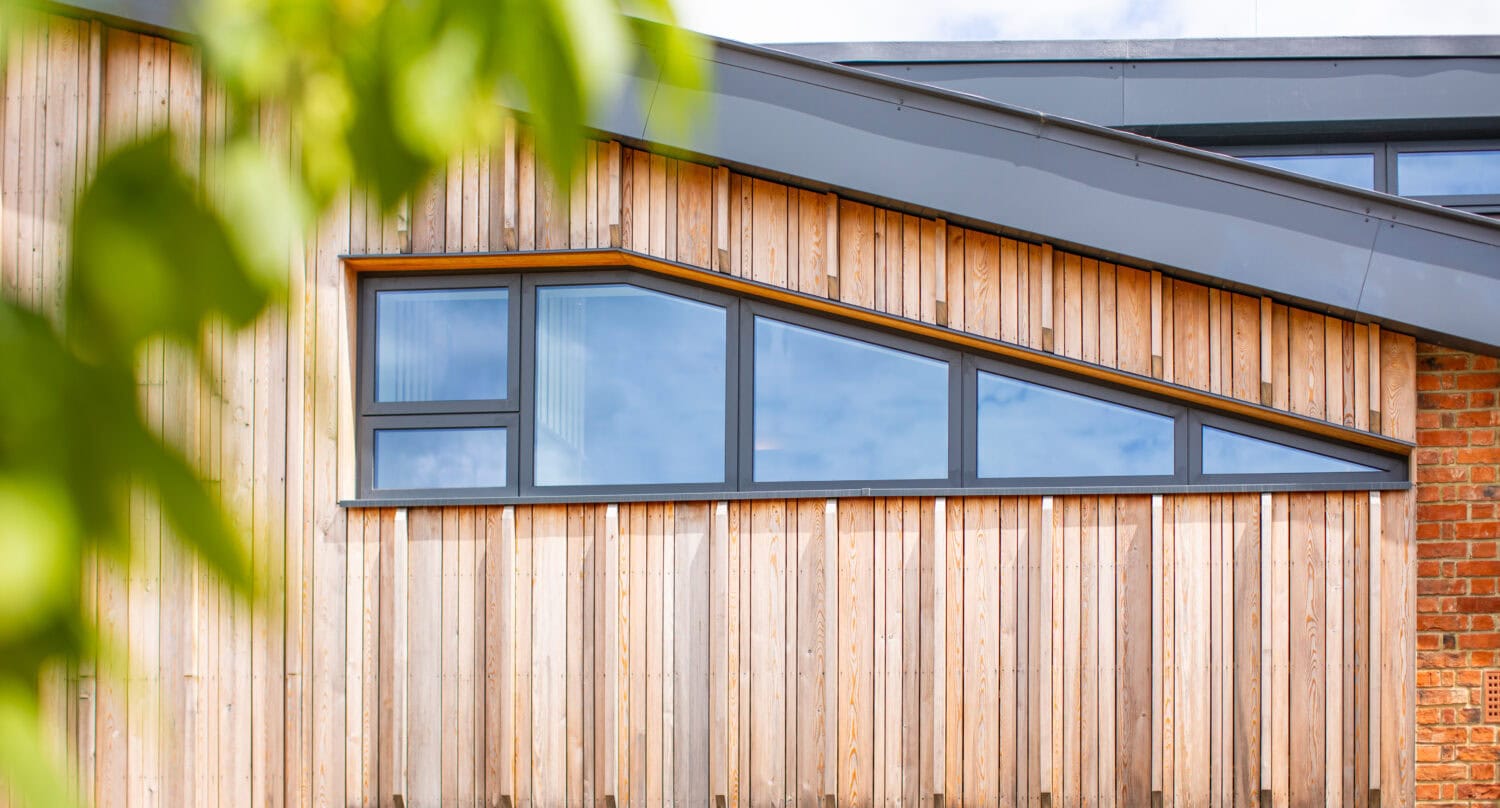 Modern building facade with light wooden panels, large rectangular and triangular windows, and part of a sloped dark roof. Bespoke glass solutions complement the design, while green leaves are blurred in the foreground.