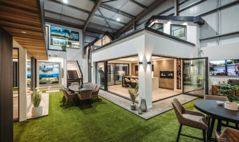 Modern indoor showroom featuring a stylish kitchen and dining area setup, large glass windows, bespoke glass solutions, artificial grass flooring, elegant furnishings, plants, and bright lighting under a high ceiling.