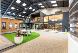 Modern indoor showroom with large windows, model buildings, a seating area on artificial grass, and bright natural lighting from skylights and glass roofing. Contemporary décor and sleek bifold doors create a spacious, open atmosphere.