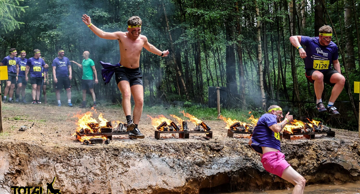 Participants in a Total Warrior obstacle race leap over fiery barriers into a muddy pit, with spectators and trees in the background. One runner lands heavily in the mud as others jump or wait to cross—no front doors or bifold doors needed here!.