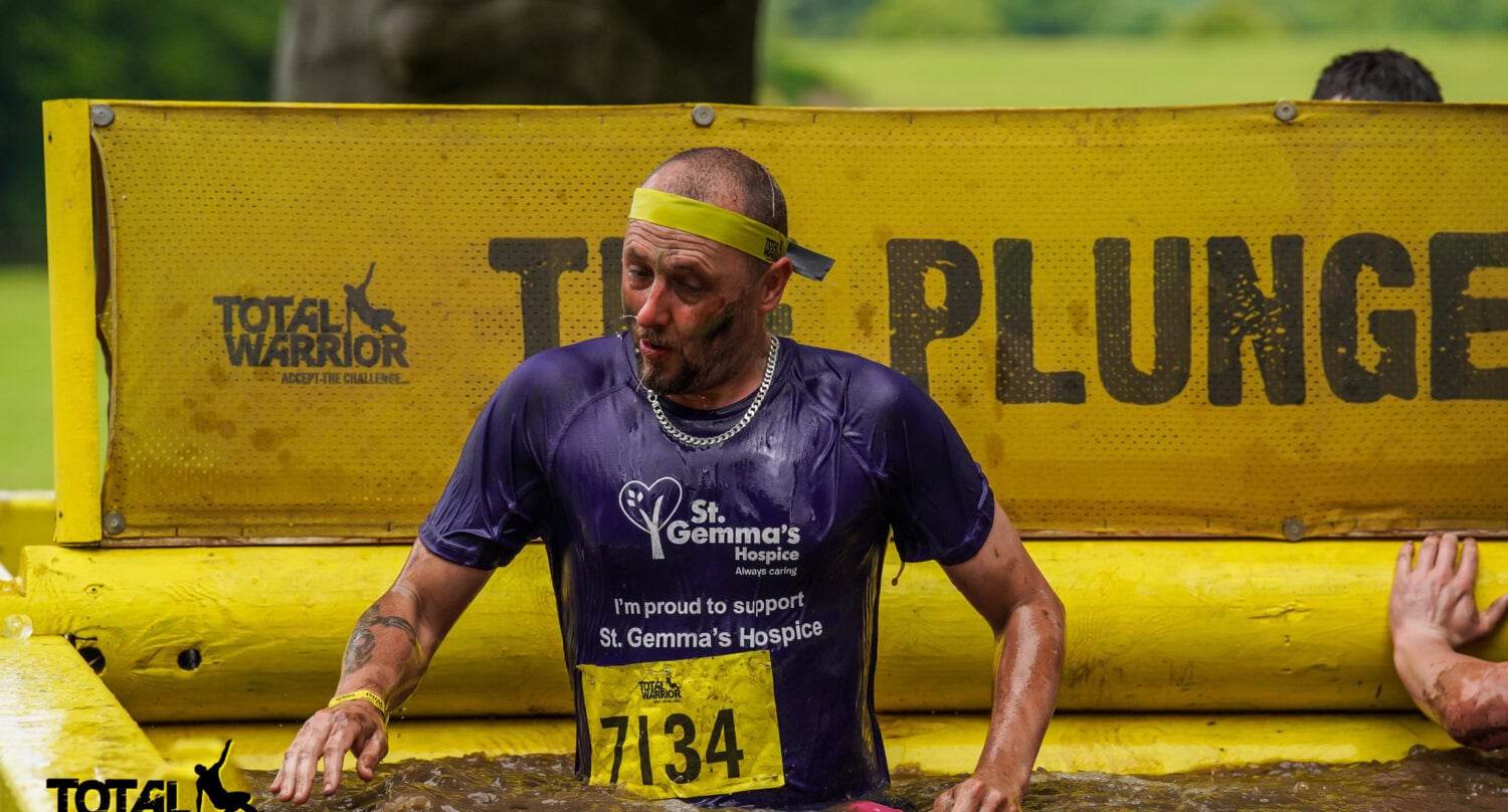 A man in a purple St. Gemma’s Hospice shirt and yellow headband emerges from a muddy obstacle at a Total Warrior event, with water up to his waist and a determined expression—ready for any challenge, from glass roofing to sliding doors.