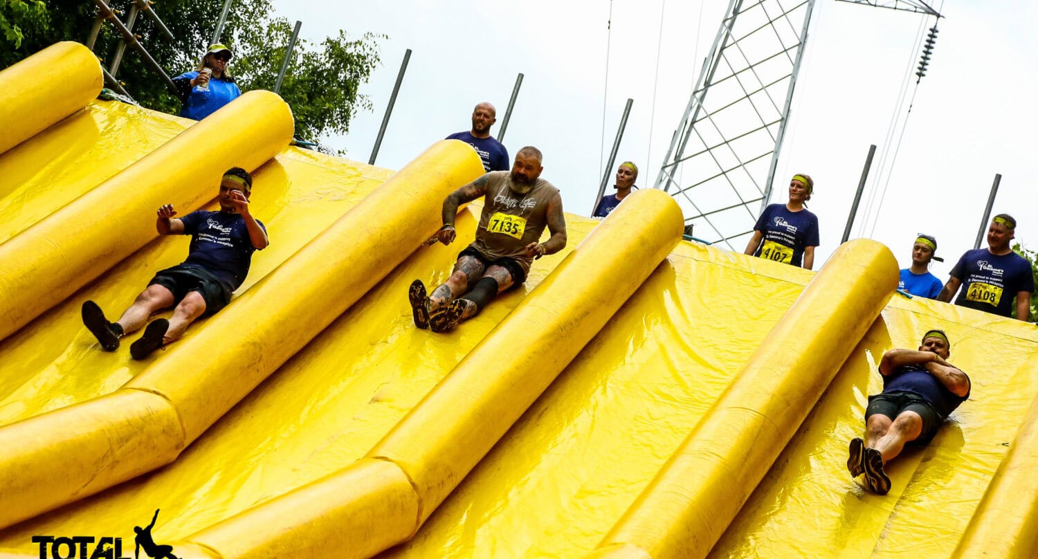 A group of people in athletic gear slide down large yellow inflatable slides during a Total Warrior obstacle course event, with onlookers at the top and trees, power lines, and glass roofing visible in the background.