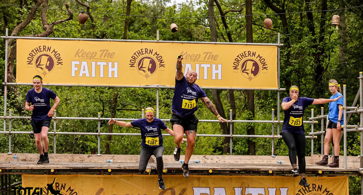 Five people in athletic gear leap off a platform labeled Leap of Faith at an outdoor obstacle event. Surrounded by trees and banners, the group’s energy rivals the excitement of glass roofing or bespoke glass solutions for any dynamic space.